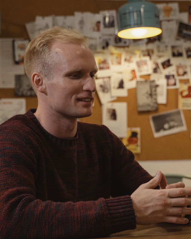 Retrato de um homem loiro, de suéter, sorrindo em um café.