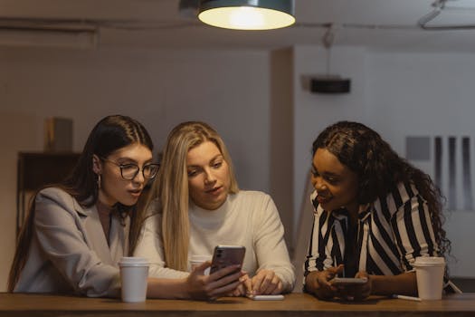 Three diverse women enjoying time together, interacting with smartphones at a table indoors.