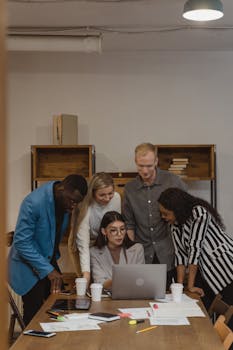A diverse group of colleagues engages in teamwork around a laptop in a modern office.