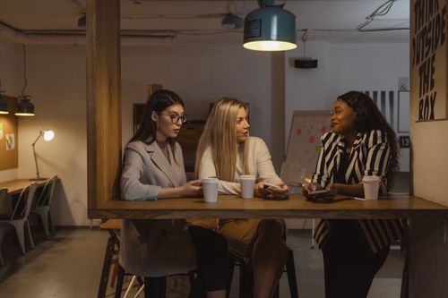 Free Three women colleagues talking over coffee at an office table, fostering communication. Stock Photo