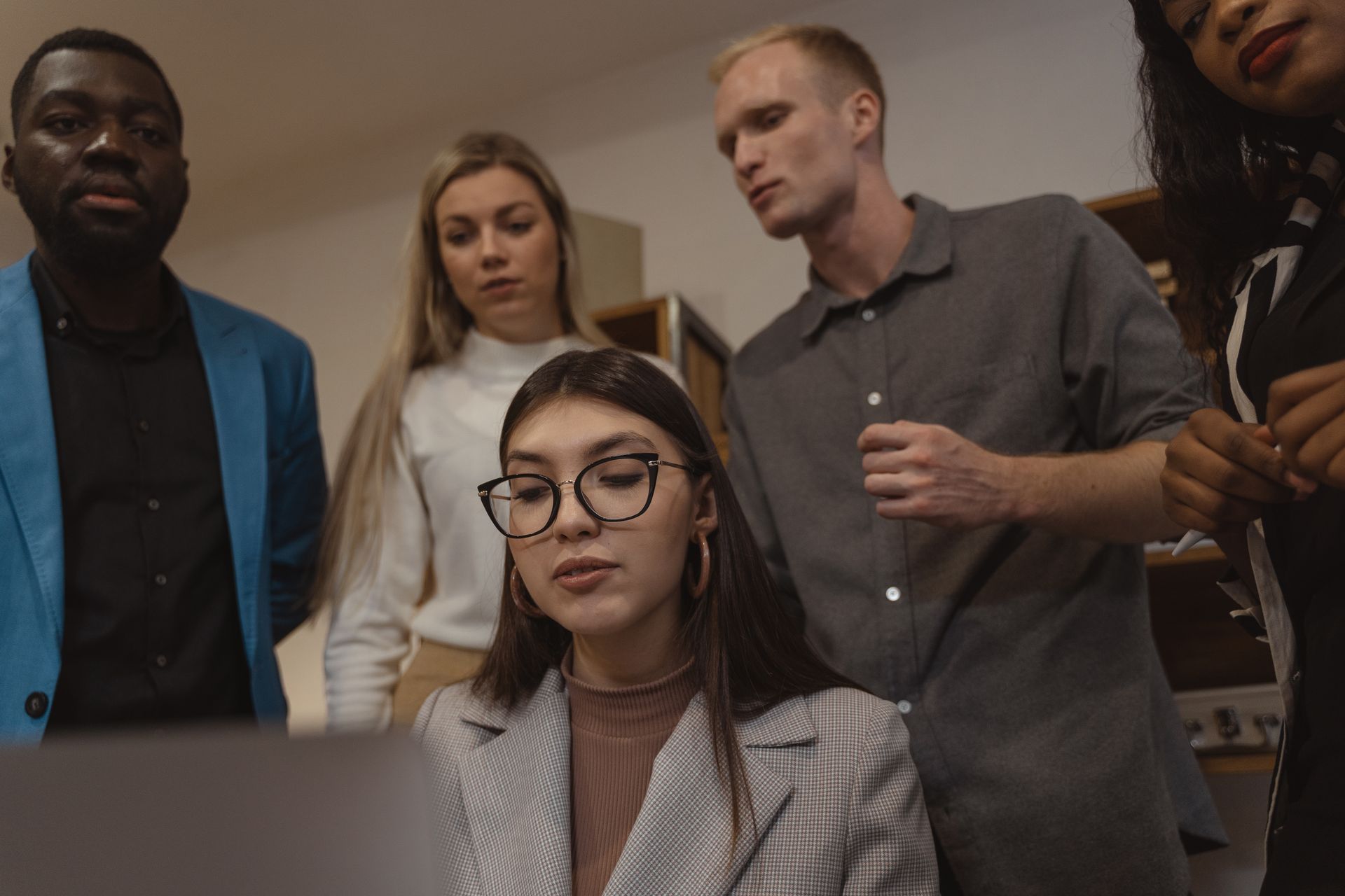 A group of professionals in a modern office setting, engaged in teamwork and collaboration around a laptop.