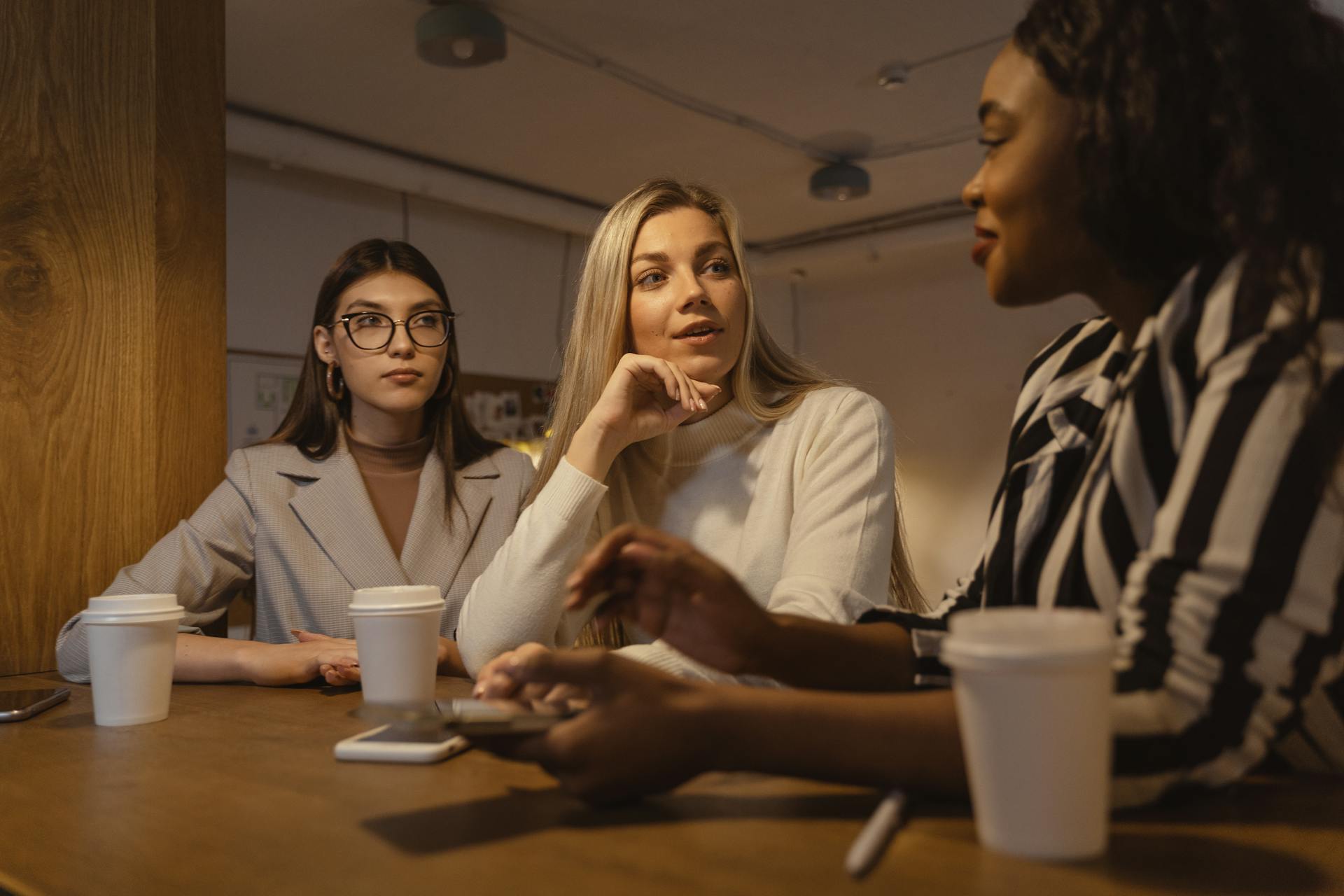 Three women engaged in a business discussion at a coffee shop, focusing on teamwork and corporate conversations.