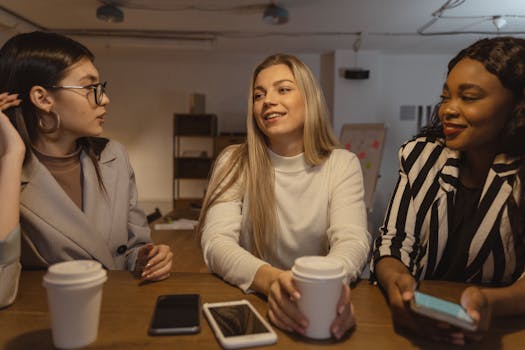 Three young women having a lively conversation over coffee in an office setting.