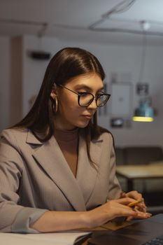 一位戴着眼镜、身着公司服装的女商人，正专注于光线昏暗的办公环境中工作