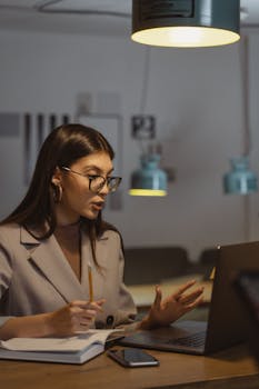 A young businesswoman working late at her desk, engaged in an online meeting with a laptop, notebook, and smartphone.