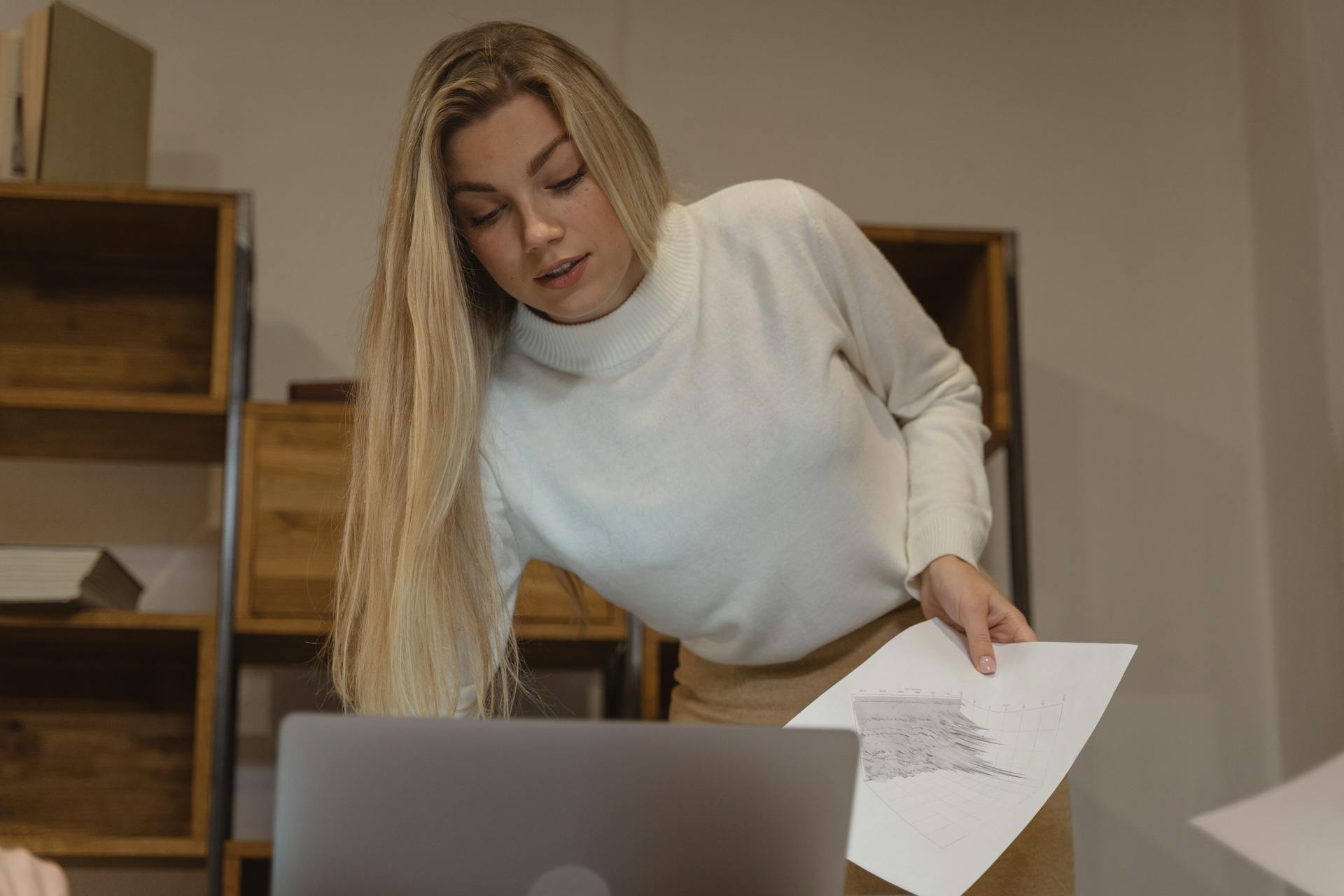 Blonde woman in a turtleneck analyzing documents by a laptop in an office setting.