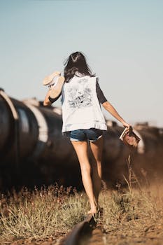 A woman in denim shorts walks along train tracks in rural Brasil, holding shoes and a book.