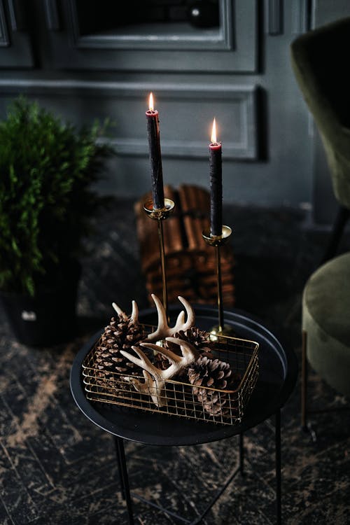 Long thin flaming candles next to pinecones and small horns in metallic box on round table on old shabby wooden floor