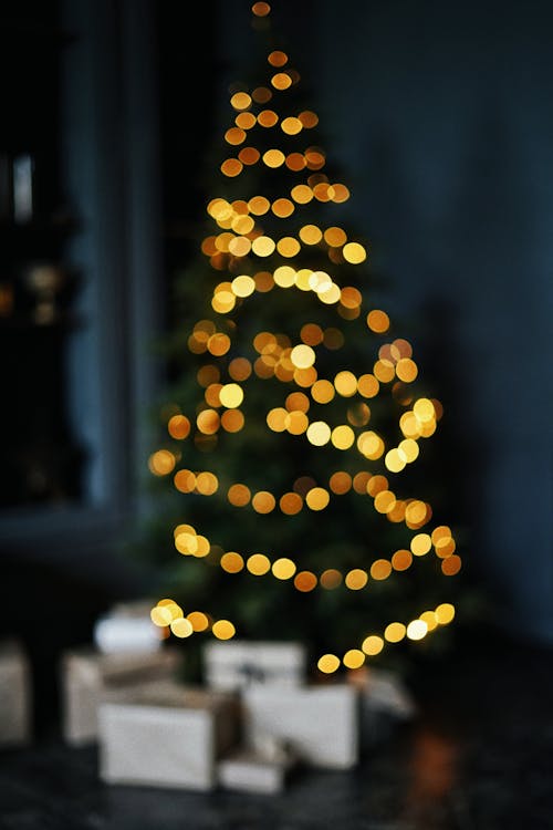 Defocused bunch of boxes under tall green Christmas tree decorated with bright yellow glowing garlands