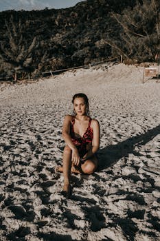 一位身材苗条、头发湿漉漉的女性穿着泳衣坐在沙滩上，看着镜头