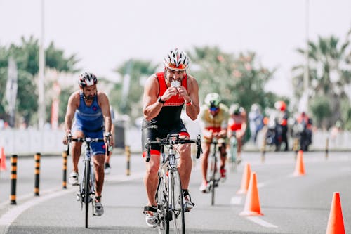 Fit male bicyclist drinking beverage while riding bicycle behind unrecognizable rivals during race on roadway in city