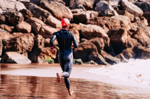 Free Back view of anonymous barefoot male athlete in diving suit jogging on sandy sea shore near stones Stock Photo