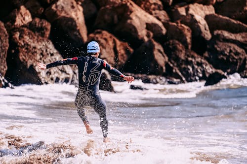Back view of unrecognizable barefoot sportsman in wet suit jumping on wavy ocean with splashing water near boulders