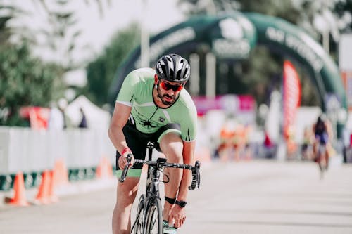 Bearded male cyclist in sportswear and helmet riding bicycle while touching sneaker on asphalt roadway during race