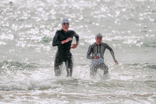 Focused young people in wet suits and swim caps running out of wavy glistening sea in sunlight