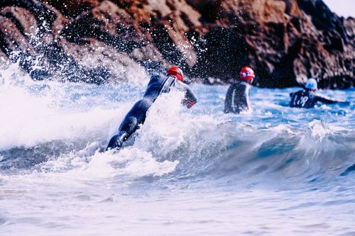 Free Group of faceless sportspeople in suit for bathing and swim caps jumping into azure wavy sea near steep mountain in sunlight Stock Photo