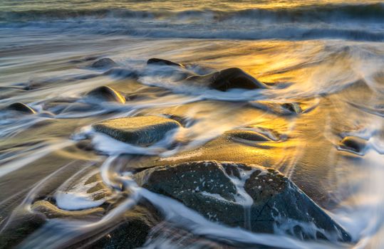 Dynamic coastal scene with waves flowing over rocks at golden sunset.