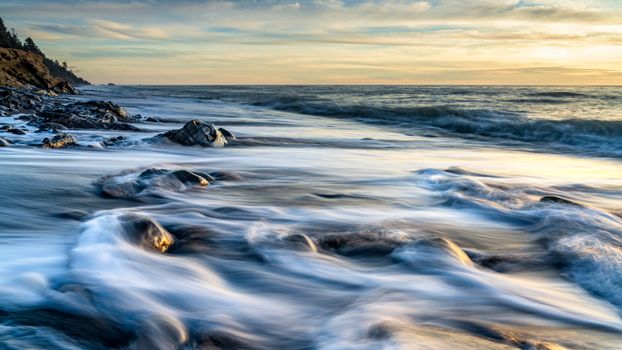 Calm waves flow over rocks at sunset on a serene ocean beach.