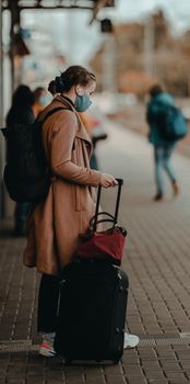 A woman in a coat and mask waits with luggage at an outdoor train station.