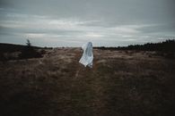 Anonymous person wearing white sheet in meadow with dried grass near forest under cloudy gloomy sky
