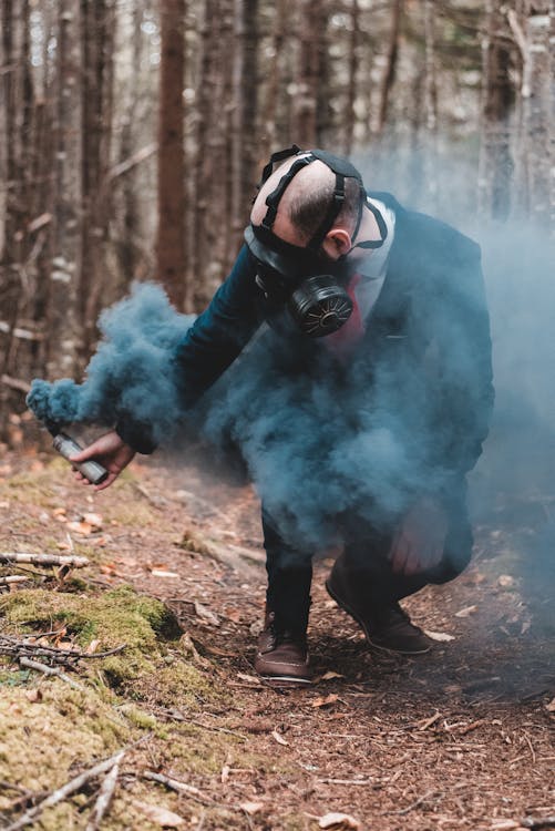 Free Confident man in protective gas mask squatting down with black color bomb in hand against trees in forest Stock Photo