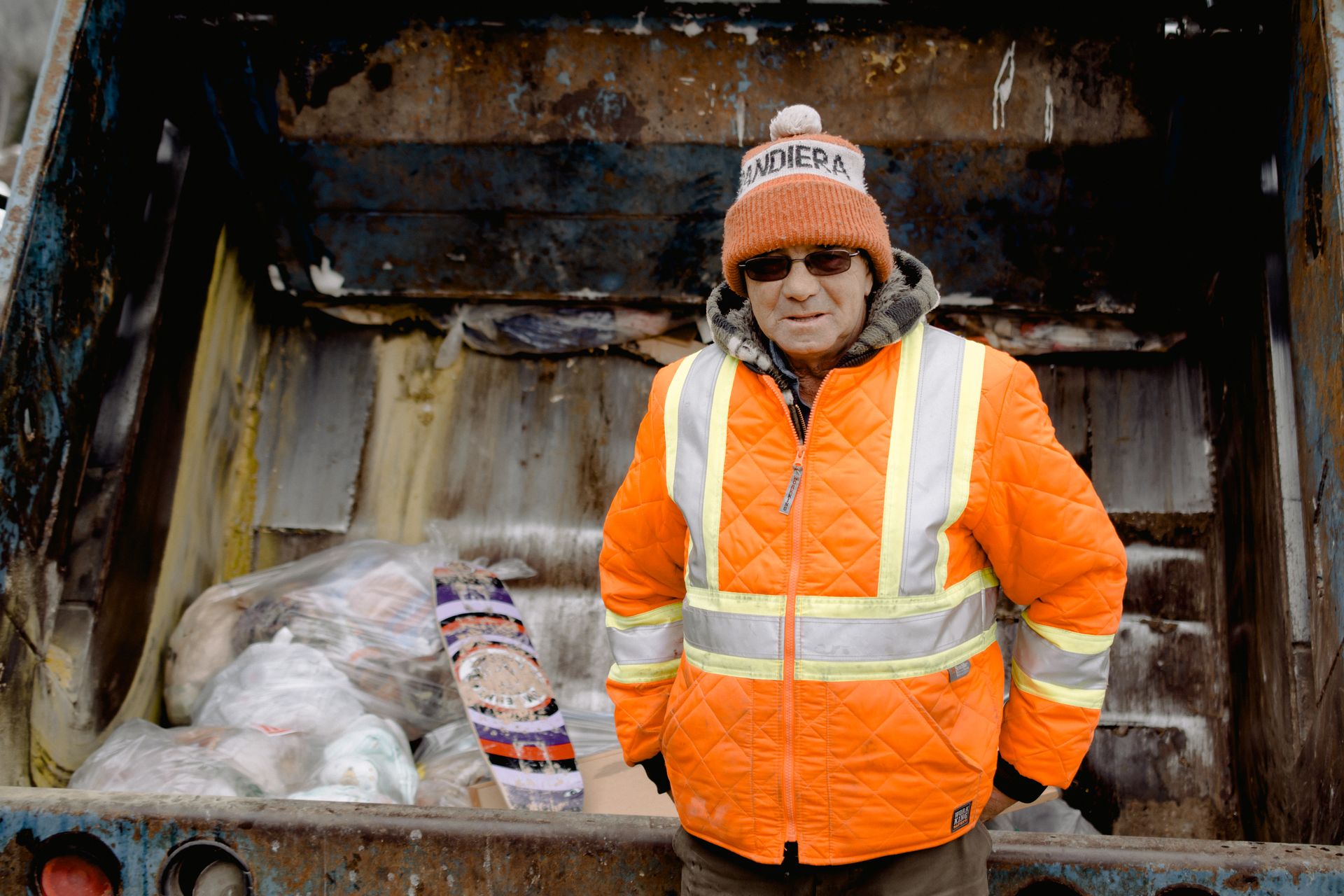 Mature dustman in colorful uniform with stripes and sunglasses standing with hands on hips near old truck with garbage