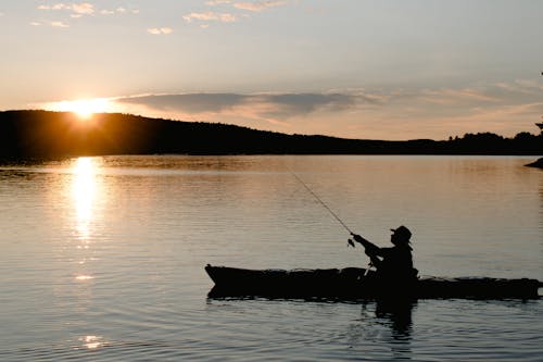 Side view of anonymous male silhouette in boat catching fish with rod on rippled river under cloudy sky with shiny sun at dusk