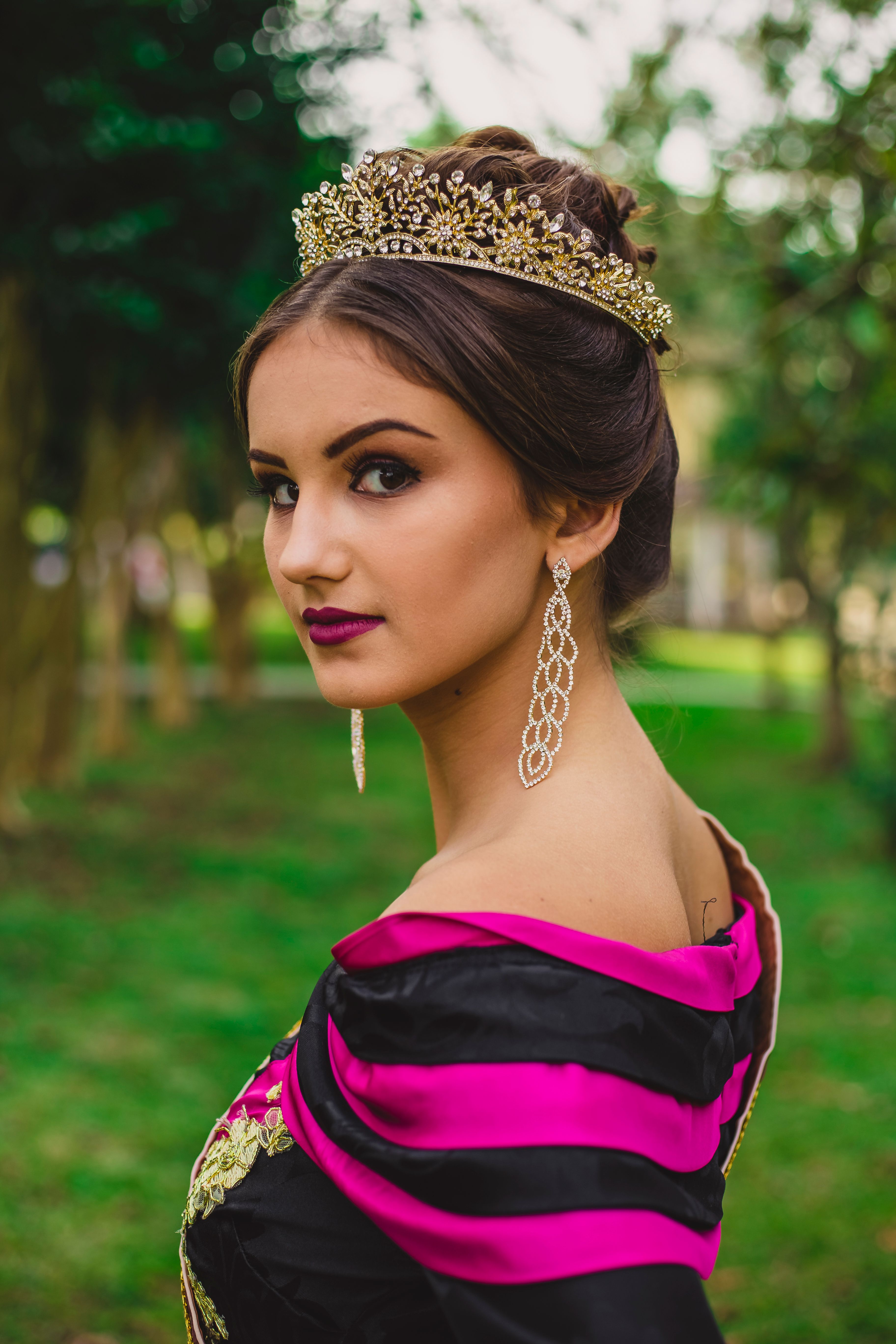 Side view of young attentive woman in crown and earrings looking at camera on lawn in park