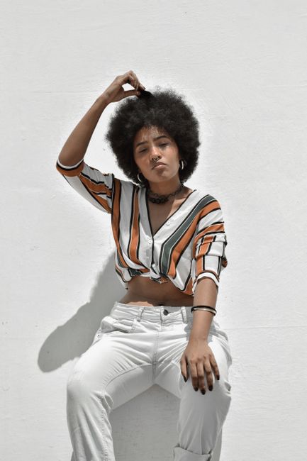 Young confident black lady with raised arm in trendy clothes combing hair on white background in sunlight