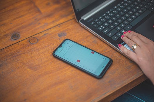 Close-up of a hand using a laptop with a smartphone showing a calendar on a wooden desk.