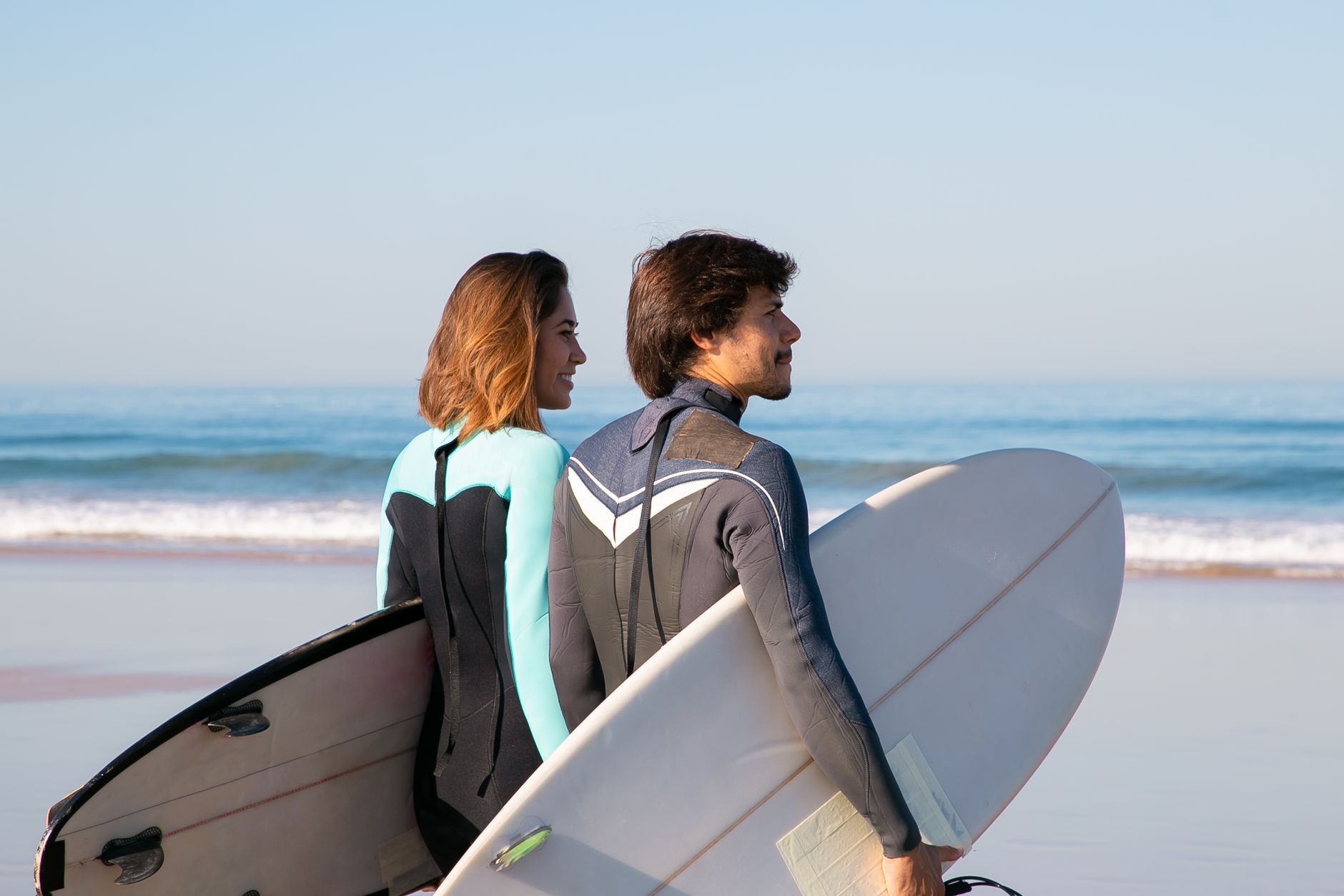 A couple in wetsuits enjoys a surfing adventure on a picturesque beach in Portugal under clear skies.