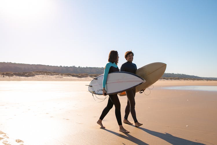 Couple Carrying Surfboards While Walking On Sand