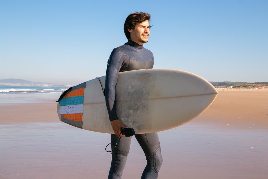 A man in a wetsuit carrying a surfboard on a sunny Portuguese beach.