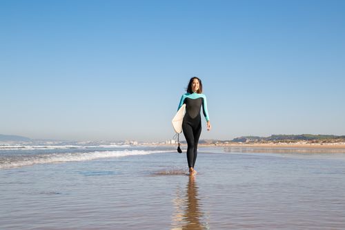 Free Asian woman with surfboard walking on a sunny beach in Portugal, perfect for summer sports and travel concepts. Stock Photo