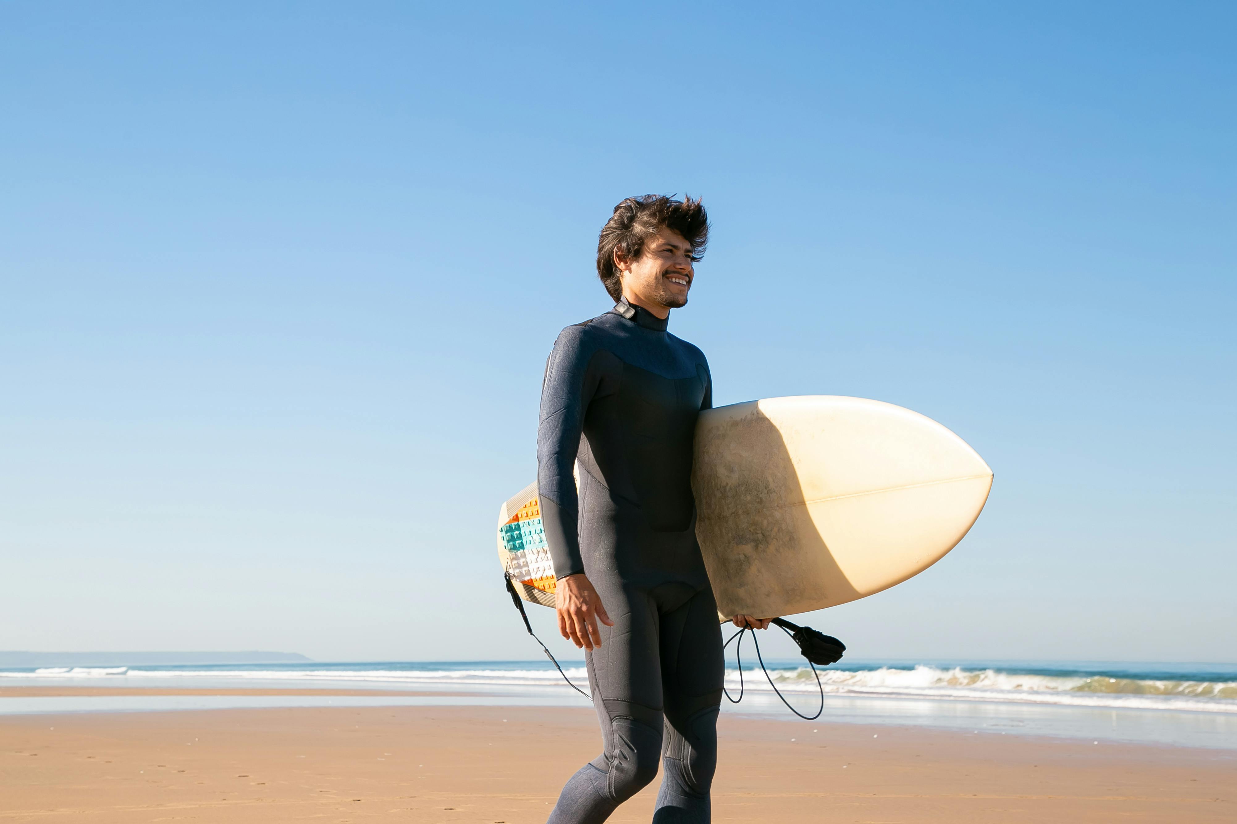 Man walking on a sunny beach in Portugal carrying a surfboard. Perfect day for surfing under blue skies.