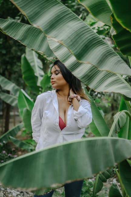 Confident young ethnic female with long dark hair in casual outfit touching neck in green tropical garden
