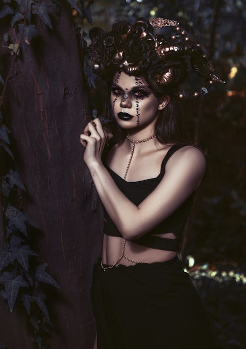 Serious young female in black outfit and spooky makeup with accessory on head standing near tree