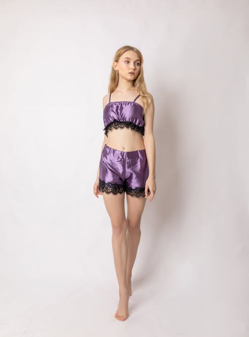 Full body of slim barefoot calm female model wearing stylish silk purple sleepwear standing in studio