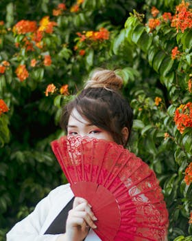 Portrait of an Asian woman with a red fan surrounded by lush orange flowers in a garden.