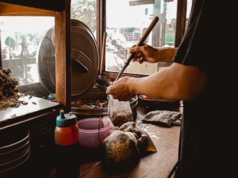 Street food vendor preparing a traditional Asian noodle dish in a local outdoor market scene.