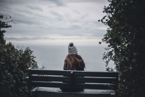 Free Woman in a beanie sits on a bench, gazing at the tranquil ocean view. Stock Photo