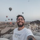 A Bearded Man in White Shirt Smiling while Taking a Selfie Near the Hot Air Balloons