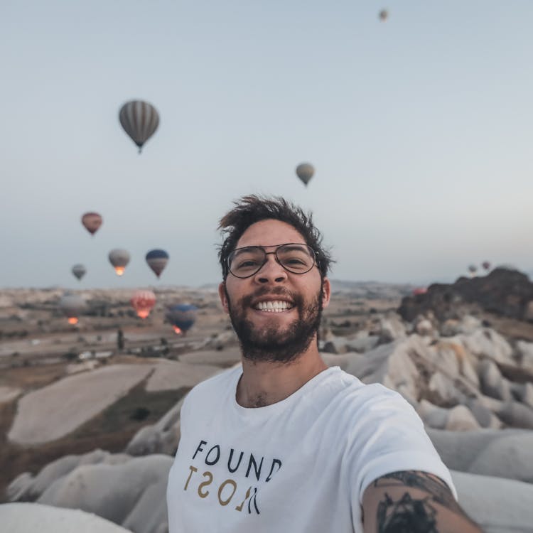 A Bearded Man In White Shirt Smiling While Taking A Selfie Near The Hot Air Balloons