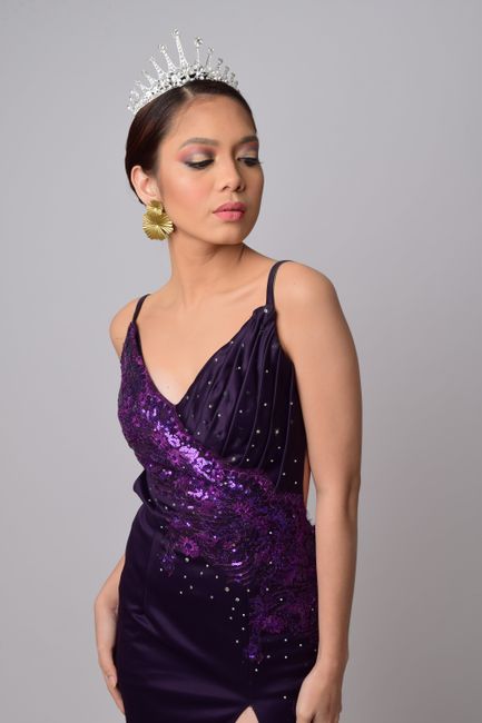 Graceful woman in a purple dress with a tiara and gold earring, posing against a gray background.