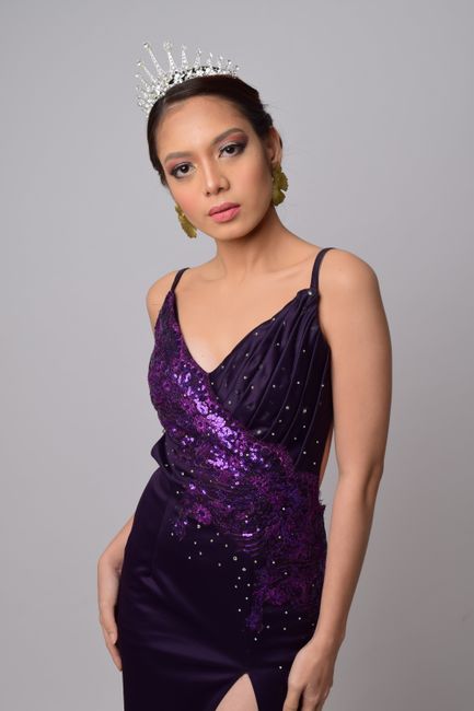 Portrait of a woman in a sequined purple gown and tiara against a gray background.