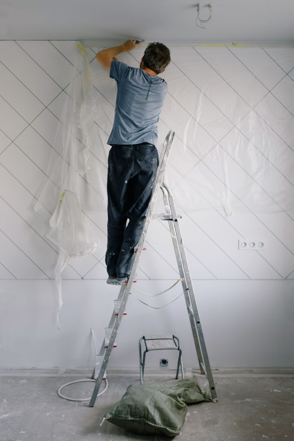 Painter on an aluminum stepladder rolling a pale wall, with plastic sheeting draped over the paneling behind