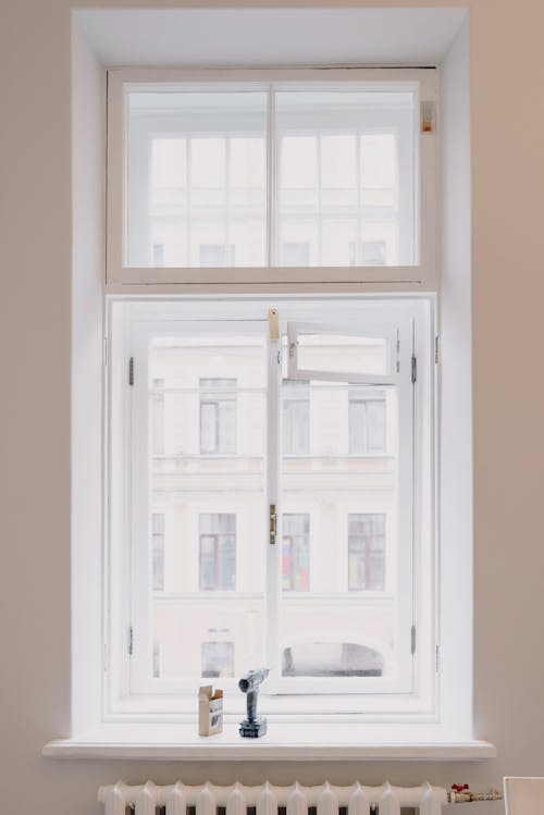 Wooden window with white frame overlooking building in city installed in light room with battery and screwdriver on windowsill in apartment