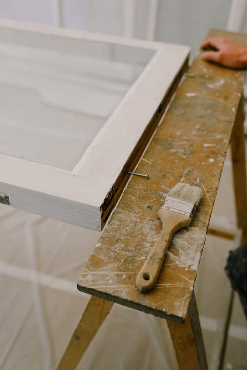 From above of anonymous craftsman standing near wooden workbench with brush and window with white frame in room during repair works