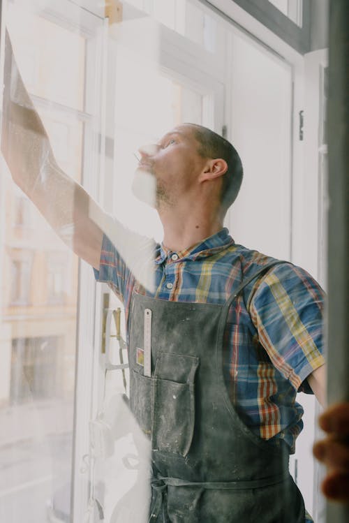 Professional focused craftsman in uniform standing with raised arm standing behind glass while assembling window in apartment during renovation works