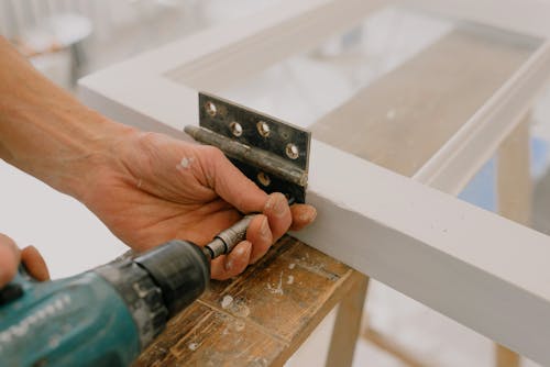 Free Crop unrecognizable worker drilling metal hinge on white window frame during house renovation Stock Photo